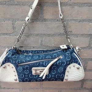 May include: A blue denim handbag with white strap and accents. The bag features a floral pattern and silver-tone hardware, including a chain strap and studded details. The bag has a zip closure and a small white tassel.