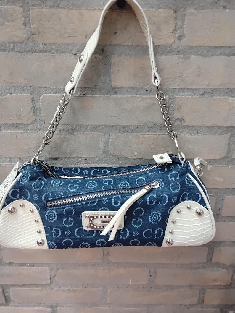 May include: A blue denim handbag with white strap and accents. The bag features a floral pattern and silver-tone hardware, including a chain strap and studded details. The bag has a zip closure and a small white tassel.