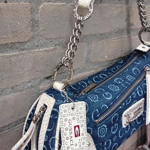 May include: A denim-style handbag with a white, textured trim and strap. The bag features silver-tone hardware, including a chain strap and zipper pulls. A tag with the brand name "Gussaci" is attached.