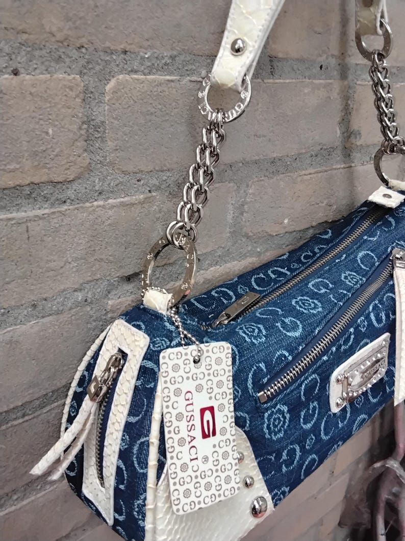 May include: A denim-style handbag with a white, textured trim and strap. The bag features silver-tone hardware, including a chain strap and zipper pulls. A tag with the brand name "Gussaci" is attached.