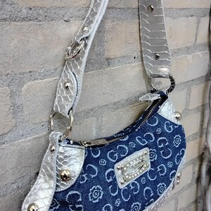 May include: A denim handbag with a silver strap and accents. The bag features a blue denim body with a repeating floral and "G" pattern. Silver hardware includes a zipper, studs, and a decorative plate with the brand name.