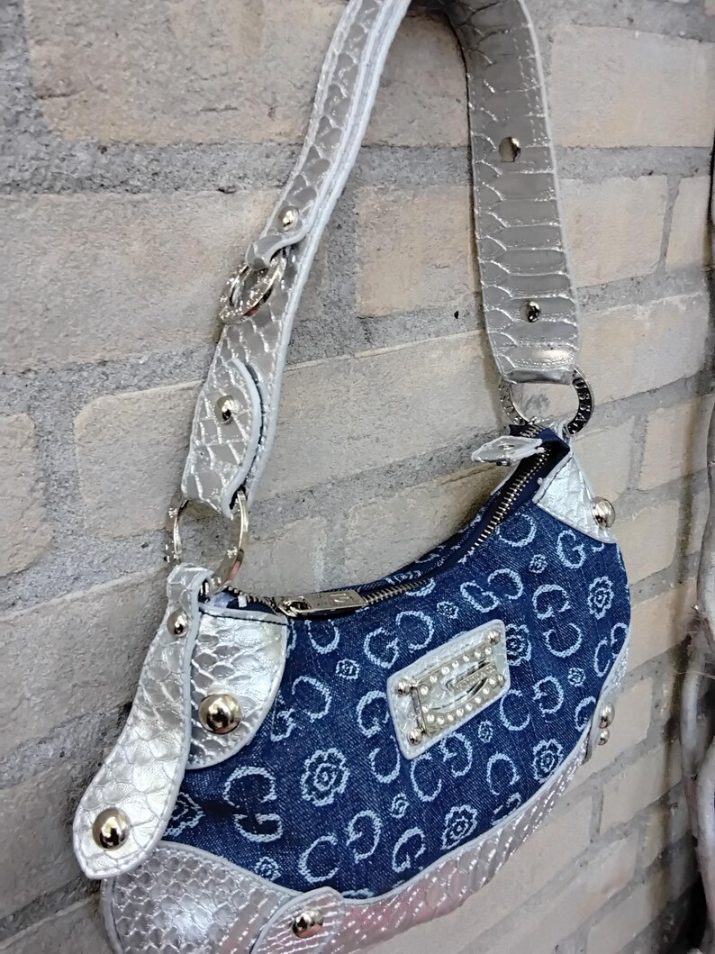 May include: A denim handbag with a silver strap and accents. The bag features a blue denim body with a repeating floral and "G" pattern. Silver hardware includes a zipper, studs, and a decorative plate with the brand name.