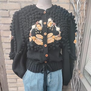 Op de afbeelding: Zwarte gebreide cardigan met een ronde hals en korte mouwen. De trui heeft een textuurontwerp met decoratieve bloemenborduursels in beige en bruin. Het heeft houten knopen en een taille met trekkoord.