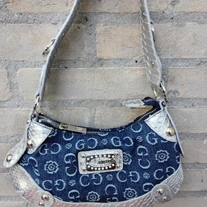 May include: A denim handbag with a silver strap and trim. The bag features a repeating pattern of the letters "G" and floral designs. A silver plaque with the brand name is centered on the front. The bag is hanging against a brick wall.