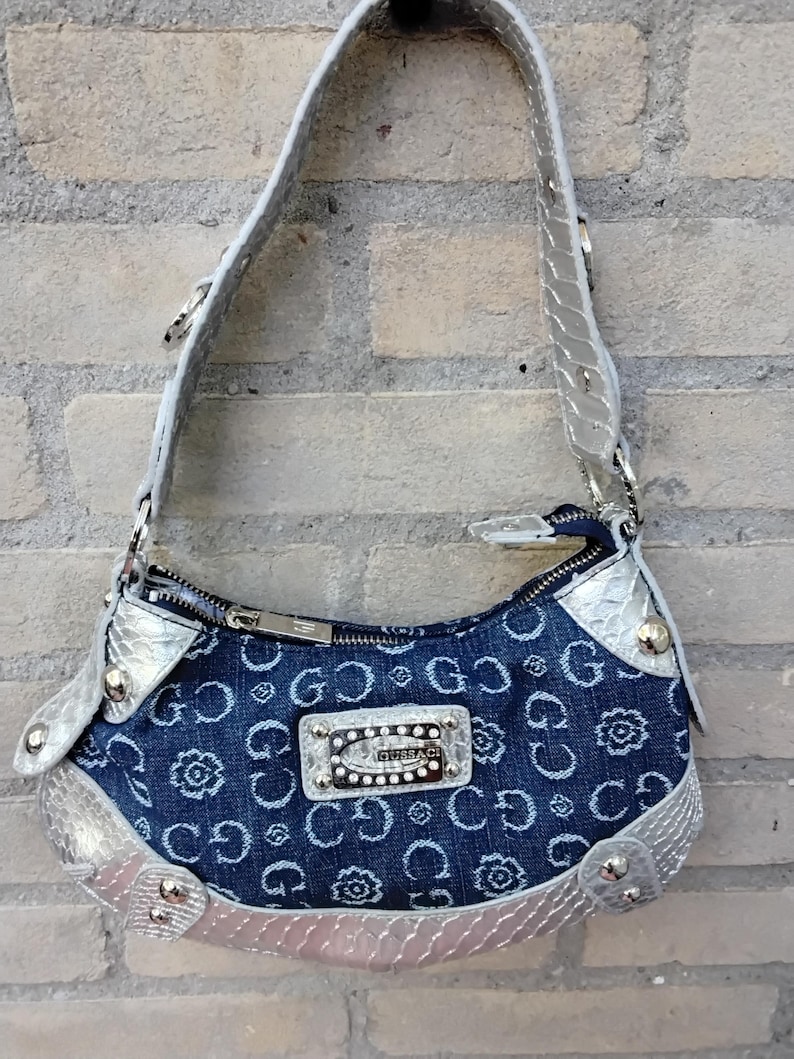 May include: A denim handbag with a silver strap and trim. The bag features a repeating pattern of the letters "G" and floral designs. A silver plaque with the brand name is centered on the front. The bag is hanging against a brick wall.