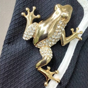Op de afbeelding: Een gouden kikker broche met heldere strass steentjes. De kikker zit op een zwarte stoffen achtergrond.