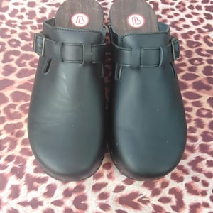 Op de afbeelding: Een paar zwarte leren klompen met een gespsluiting. De klompen hebben een houten basis en een rood hartlogo bovenop.