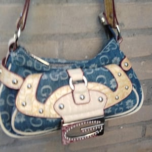 May include: A denim shoulder bag with a repeating "G" pattern and floral accents. The bag features cream-coloured leather accents, a silver buckle, and a matching strap. The bag is approximately 25 cm wide.