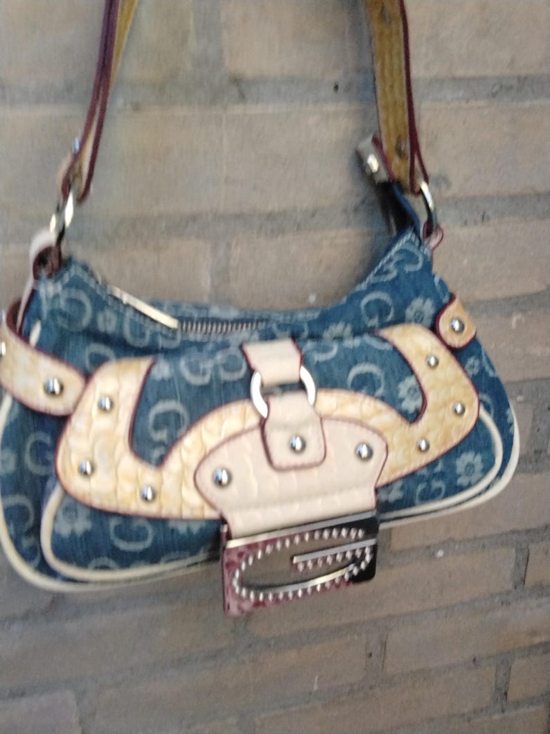 May include: A denim shoulder bag with a repeating "G" pattern and floral accents. The bag features cream-coloured leather accents, a silver buckle, and a matching strap. The bag is approximately 25 cm wide.