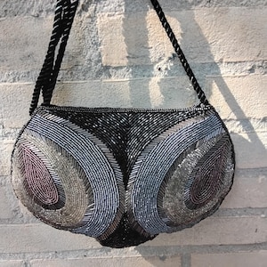 May include: A black beaded handbag with a rounded shape and a black rope strap. The bag features a pattern of silver, gray, and purple beads. The bag is hanging against a light-colored brick wall.