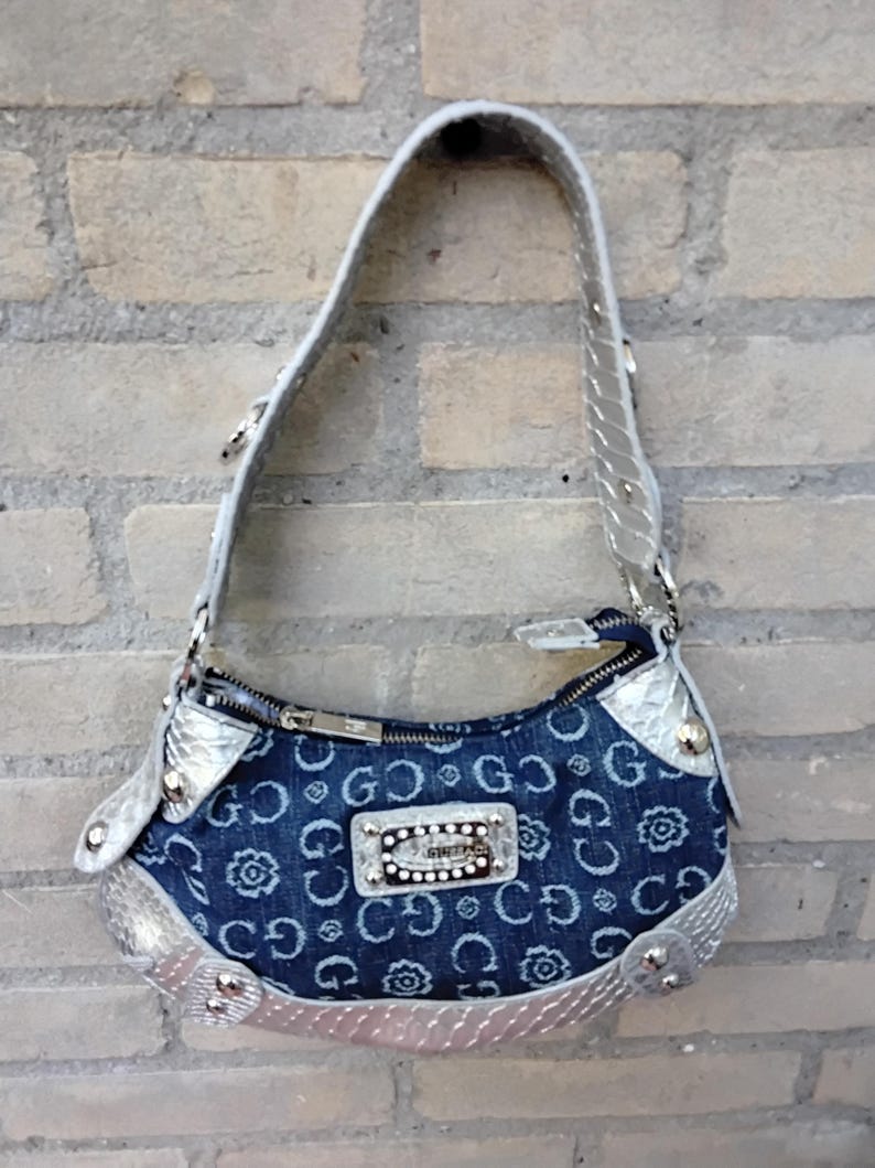 May include: A denim handbag with a silver handle and trim. The bag features a repeating pattern of the letters "G" and floral designs. A small rectangular silver plaque with the word "GUESS" is centered on the front.