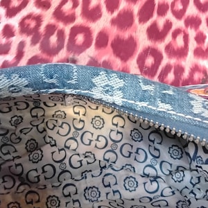 May include: Close-up of a denim handbag with a zip closure. The interior lining has a repeating pattern of the letter "G" and floral designs. The background is a pink leopard print.