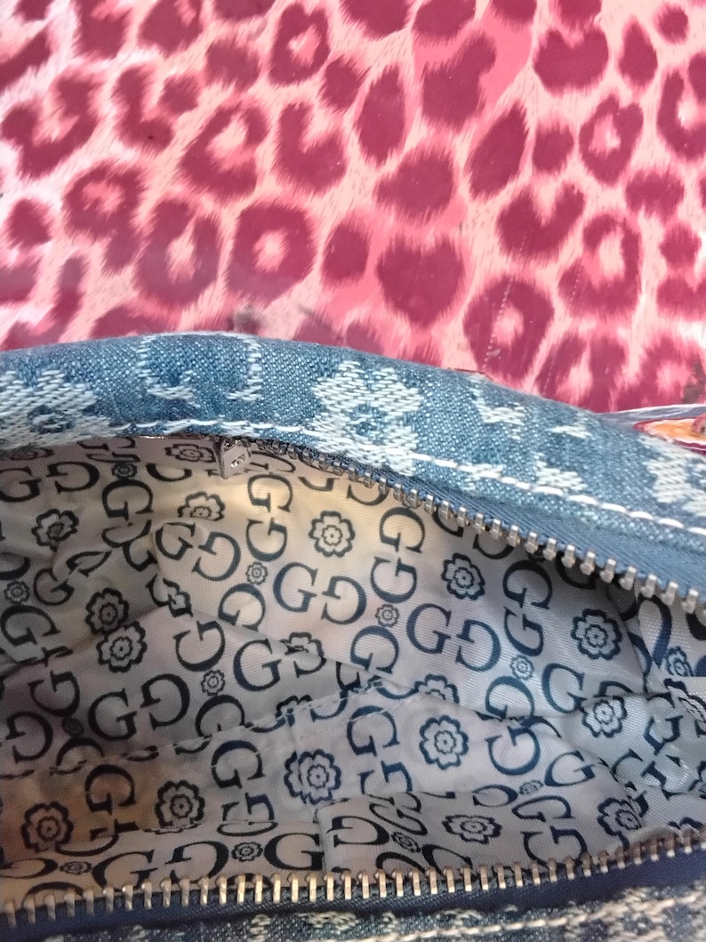 May include: Close-up of a denim handbag with a zip closure. The interior lining has a repeating pattern of the letter "G" and floral designs. The background is a pink leopard print.