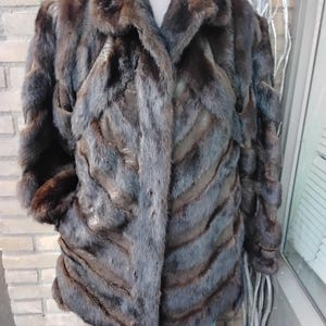 Op de afbeelding: Een donkerbruine bontjas met een reverskraag en een visgraatpatroon. De jas heeft lange mouwen en lijkt gemaakt te zijn van een zacht, luxueus materiaal. De jas wordt op een mannequin gepresenteerd.