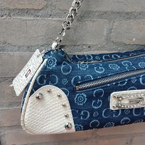 May include: A blue denim handbag with a silver chain strap. The bag has a white textured section with silver studs and a zip closure. The denim is patterned with a floral design. A white tag with text hangs from the chain.