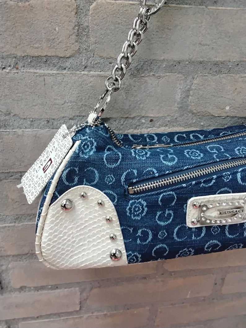 May include: A blue denim handbag with a silver chain strap. The bag has a white textured section with silver studs and a zip closure. The denim is patterned with a floral design. A white tag with text hangs from the chain.