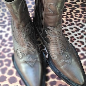 May include: A pair of brown leather cowboy boots with pointed toes and intricate stitching. The boots have a Western style and are made of a soft, supple leather.