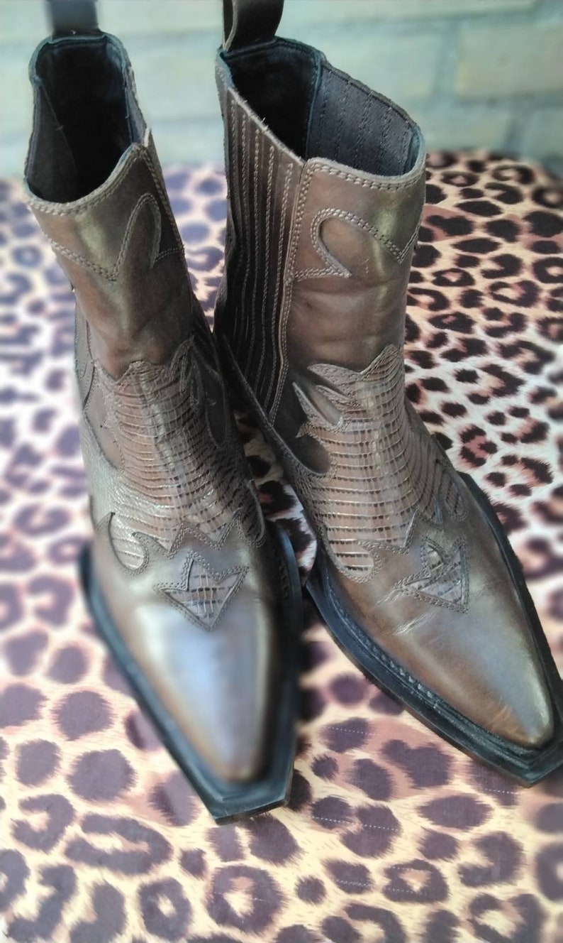 May include: A pair of brown leather cowboy boots with pointed toes and intricate stitching. The boots have a Western style and are made of a soft, supple leather.