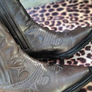 May include: A pair of brown leather cowboy boots with intricate stitching and a pointed toe. The boots have a textured pattern on the leather and a black sole.