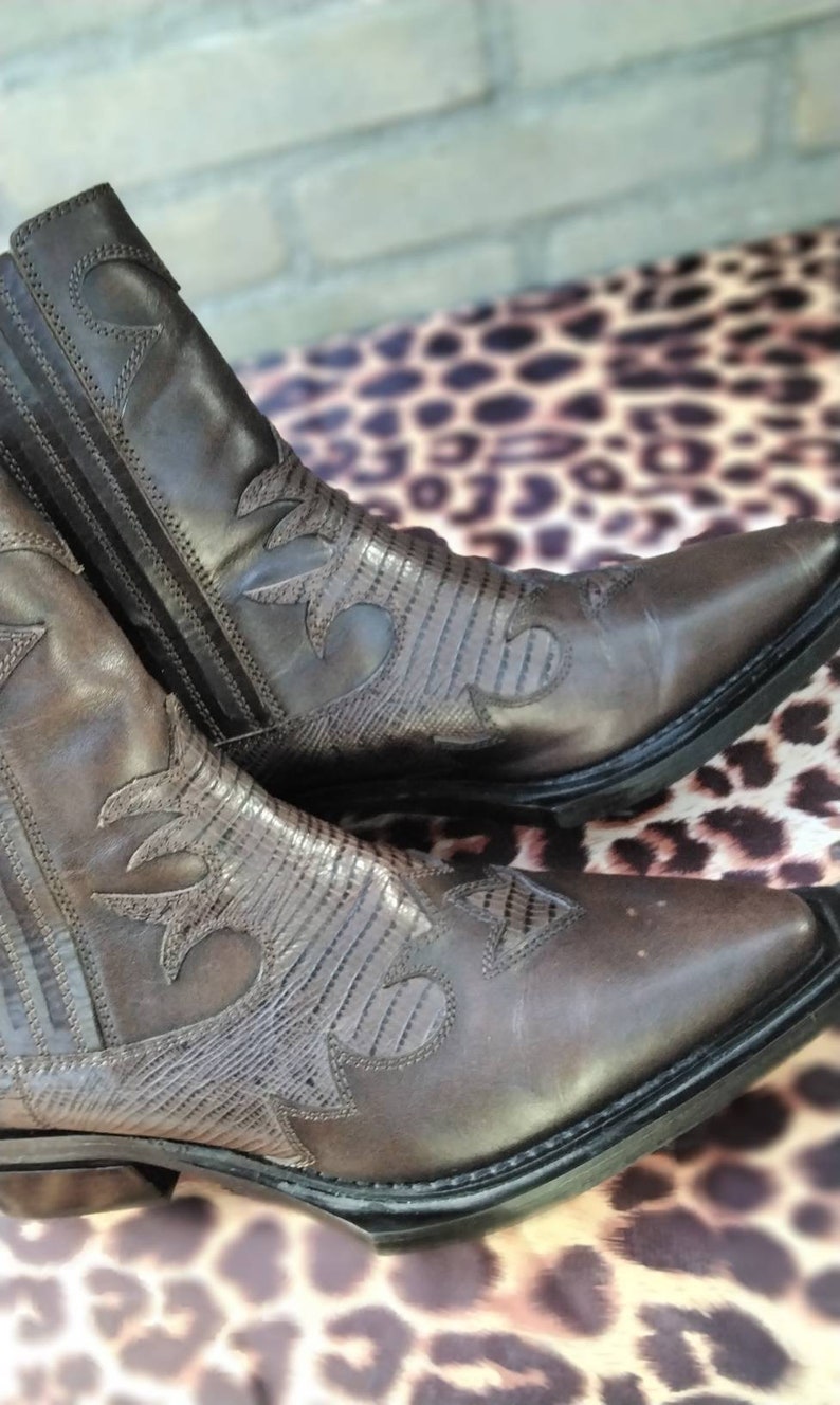 May include: A pair of brown leather cowboy boots with intricate stitching and a pointed toe. The boots have a textured pattern on the leather and a black sole.