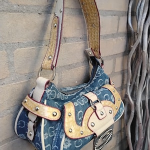 May include: A denim handbag with a repeating white "G" pattern. The bag features cream-coloured accents, a snakeskin-textured strap, and silver-toned hardware. A small, black tag with rhinestones hangs from the front.