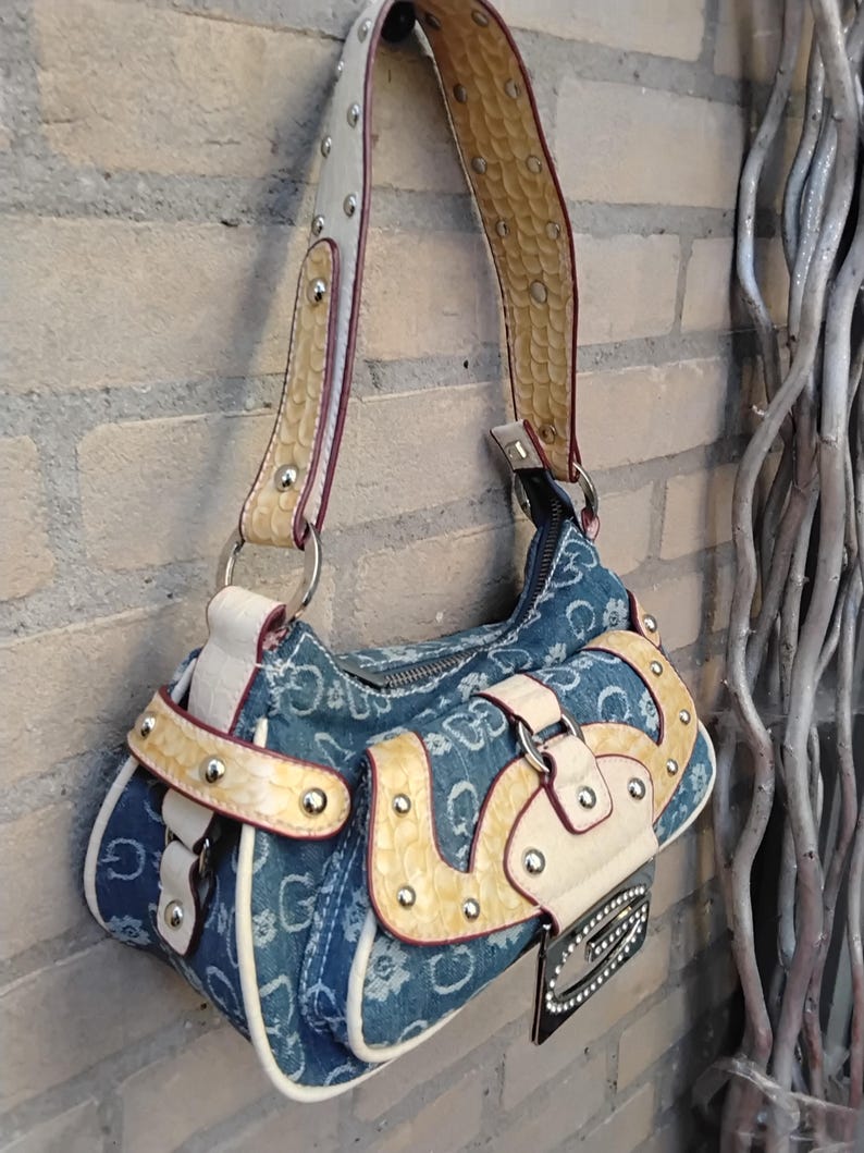 May include: A denim handbag with a repeating white "G" pattern. The bag features cream-coloured accents, a snakeskin-textured strap, and silver-toned hardware. A small, black tag with rhinestones hangs from the front.