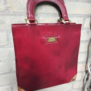 May include: A square-shaped deep red handbag with a glossy finish, featuring a rounded top handle, gold-toned metal hardware, and a rectangular gold-toned emblem on the front. The bottom corners are reinforced with gold-toned metal corner guards.