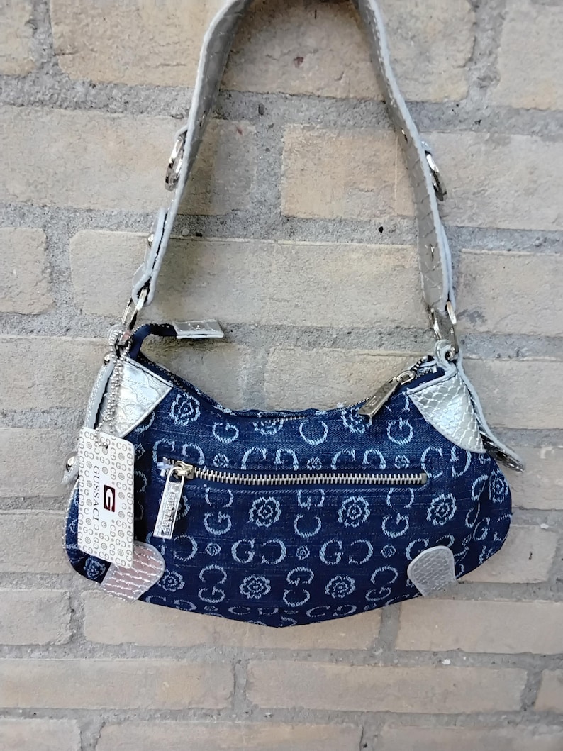 May include: A denim shoulder bag with a silver strap and accents. The bag features a repeating pattern of white floral and "G" designs. A silver zip and a tag with the brand name are visible.