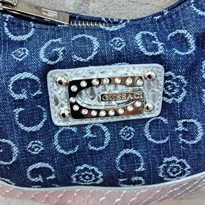 May include: A denim handbag with a repeating light blue pattern. The bag features a silver zip and a rectangular emblem with the brand name "GUSSACI" and embedded rhinestones. The bottom edge has a pink, textured trim.