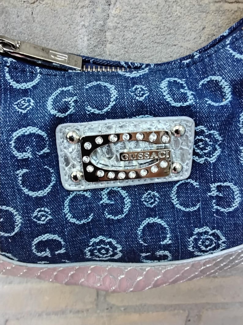 May include: A denim handbag with a repeating light blue pattern. The bag features a silver zip and a rectangular emblem with the brand name "GUSSACI" and embedded rhinestones. The bottom edge has a pink, textured trim.