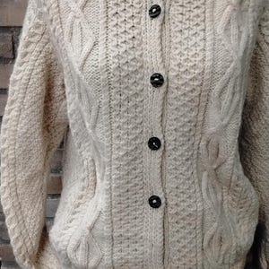 Op de afbeelding: Crèmekleurig vest met knopen en een textuurgebreid patroon. De trui heeft kabelgebreide details op de mouwen en voorpanelen, en zwarte knopen. De algehele stijl is klassiek en gezellig.