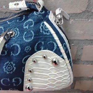May include: A denim handbag with a repeating pattern and white accents. The bag features a zip closure, silver hardware, and a white, textured panel adorned with silver studs. The bag's design incorporates a mix of textures and colours.