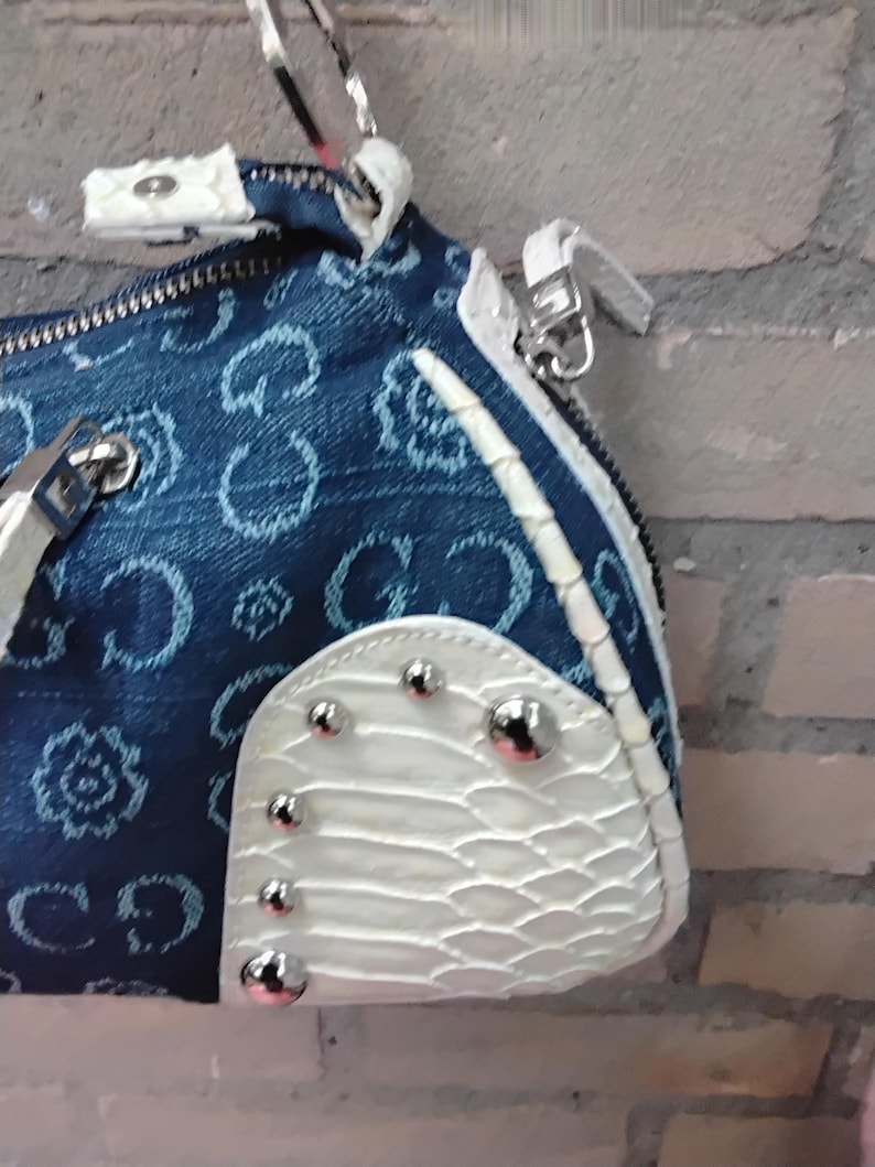 May include: A denim handbag with a repeating pattern and white accents. The bag features a zip closure, silver hardware, and a white, textured panel adorned with silver studs. The bag's design incorporates a mix of textures and colours.