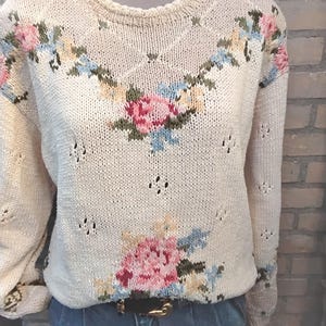 Op de afbeelding: Crèmekleurige gebreide trui met bloemenborduurwerk in roze, blauw en geel. De trui heeft een ronde hals, lange mouwen en kleine decoratieve uitsparingen. Een zwarte riem met een gouden gesp is zichtbaar.