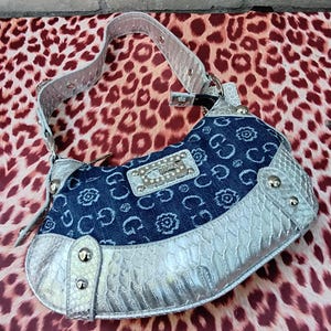 May include: A denim and silver handbag with a shoulder strap. The bag features a blue denim body with a floral and "G" pattern, silver trim, and a silver strap. The bag has a rectangular silver plate with the brand name "GUESS" on it.