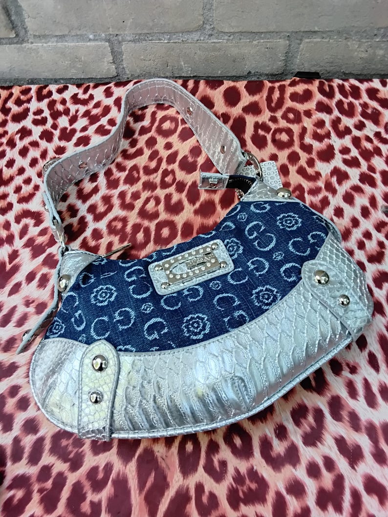 May include: A denim and silver handbag with a shoulder strap. The bag features a blue denim body with a floral and "G" pattern, silver trim, and a silver strap. The bag has a rectangular silver plate with the brand name "GUESS" on it.