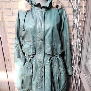 Könnte beinhalten: Türkisgrüne Lederjacke mit Pelzkapuze. Die Jacke hat eine Knopfleiste, Taschen und eine Taille mit Kordelzug. Der Pelzbesatz ist hellbraun.