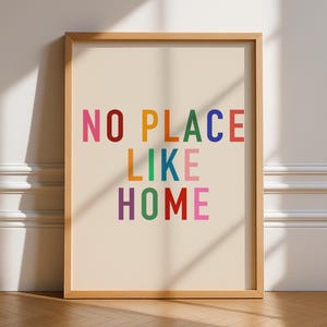 May include: A framed print featuring the phrase "NO PLACE LIKE HOME" in vibrant, multi-coloured block letters. The text is set against a cream backdrop, within a light brown wooden frame. The print is suitable for home decor.