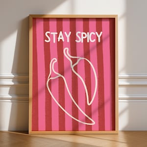May include: A framed art print with a pink and red striped background and two white chilli pepper outlines. The words "STAY SPICY" are written in white at the top. The frame is a light brown colour.