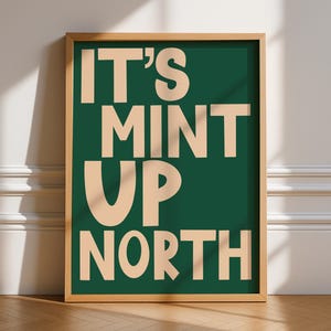 May include: A framed print with the words "IT'S MINT UP NORTH" in a beige font on a dark green background. The frame is light brown. The print is displayed on a white wall with a wooden floor.