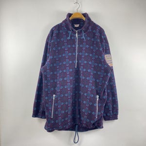 May include: A purple fleece pullover with a blue geometric pattern. The jacket features a half-zip, two zipped side pockets, and a drawstring hem. A small patch is sewn on the right sleeve.
