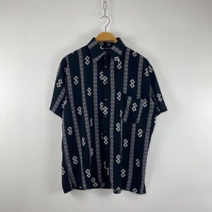 Peut inclure: Chemise noire à manches courtes avec un motif géométrique blanc. La chemise a un col, une poche poitrine et est suspendue à un cintre en bois.