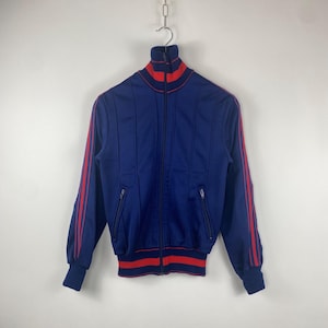 Könnte beinhalten: Vintage marineblaue Trainingsjacke mit roten Streifen auf den Ärmeln und am Kragen. Die Jacke hat einen durchgehenden Reißverschluss, zwei Vordertaschen sowie einen gerippten Saum und Bündchen. Die Jacke hängt an einer Metallkette.