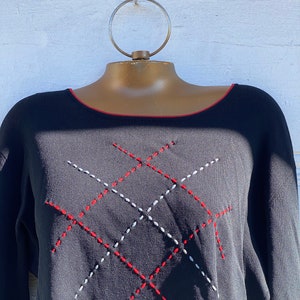 May include: Black long-sleeve sweater with a red trim around the neckline. The sweater features a stitched diamond pattern in red and white. The garment is displayed on a mannequin with a gold-colored neck and a circular hanger.