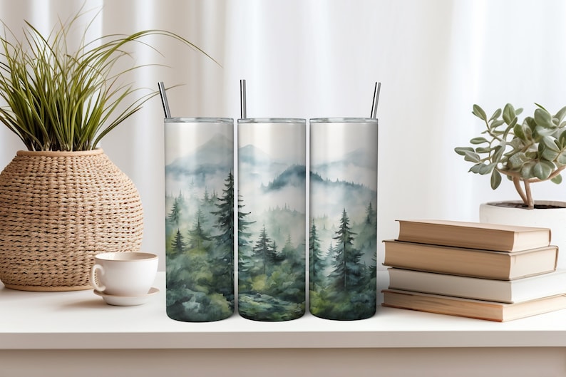 K&ouml;nnte beinhalten: Drei Edelstahlbecher mit einem Aquarelldesign eines nebeligen Waldes mit gr&uuml;nen B&auml;umen. Die Becher stehen auf einer wei&szlig;en Oberfl&auml;che mit einer Pflanze und B&uuml;chern im Hintergrund.
