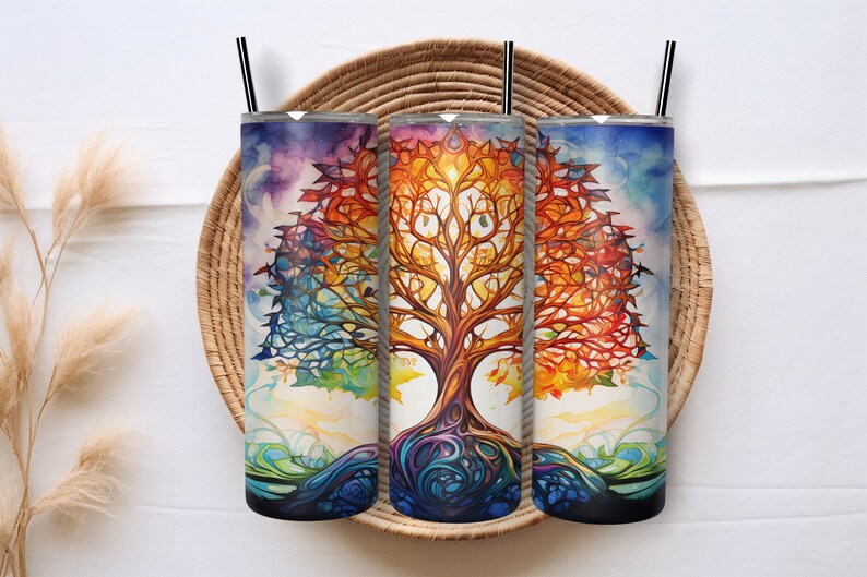 May include: Three stainless steel tumblers with a colorful watercolor tree of life design. The artwork features a vibrant spectrum of colors, including blues, oranges, and greens. Each tumbler has a black straw and a silver lid. The tumblers are displayed on a woven tray.