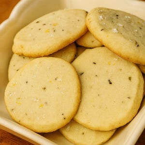 May include: A close-up shot of several round, golden-brown cookies in a cream-colored dish. The cookies have a slightly rough texture and are speckled with dark and yellow flecks. Some cookies have visible sugar crystals on top. The cookies are arranged in a pile.
