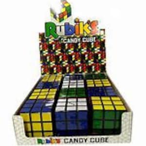 Può includere: Un espositore di cubetti di caramelle Rubik's, una caramella di fantasia a forma di cubo di Rubik. I cubetti sono neri con quadrati colorati su ogni lato. L'espositore è in cartone e riporta la scritta "Rubik's Candy Cube".