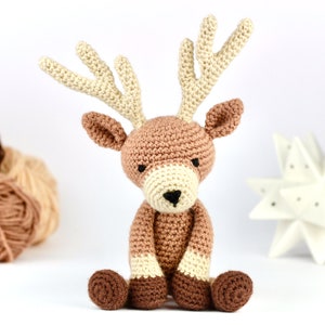 May include: A crocheted reindeer toy with brown and cream colored yarn. The reindeer is sitting with its legs crossed and has a brown nose.