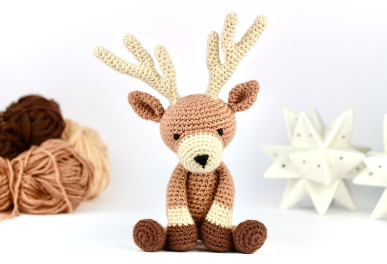 May include: A crocheted reindeer toy with brown and cream colored yarn. The reindeer is sitting with its legs crossed and has a brown nose.